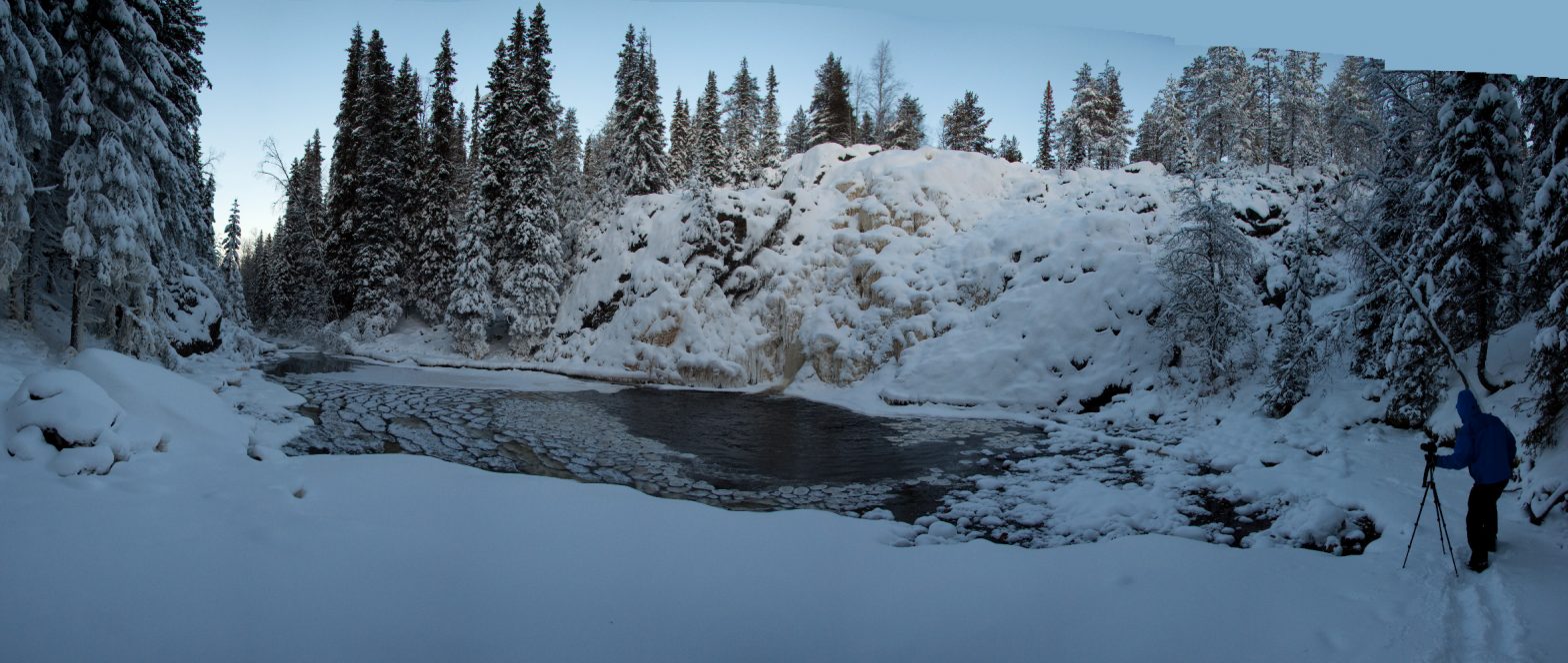 Panoraamassa jäätynyt vesiputous, sula suvanto ja kuusimetsään virtaava tummavetinen joki. Suvannon oikeassa reunassa valokuvaaja.