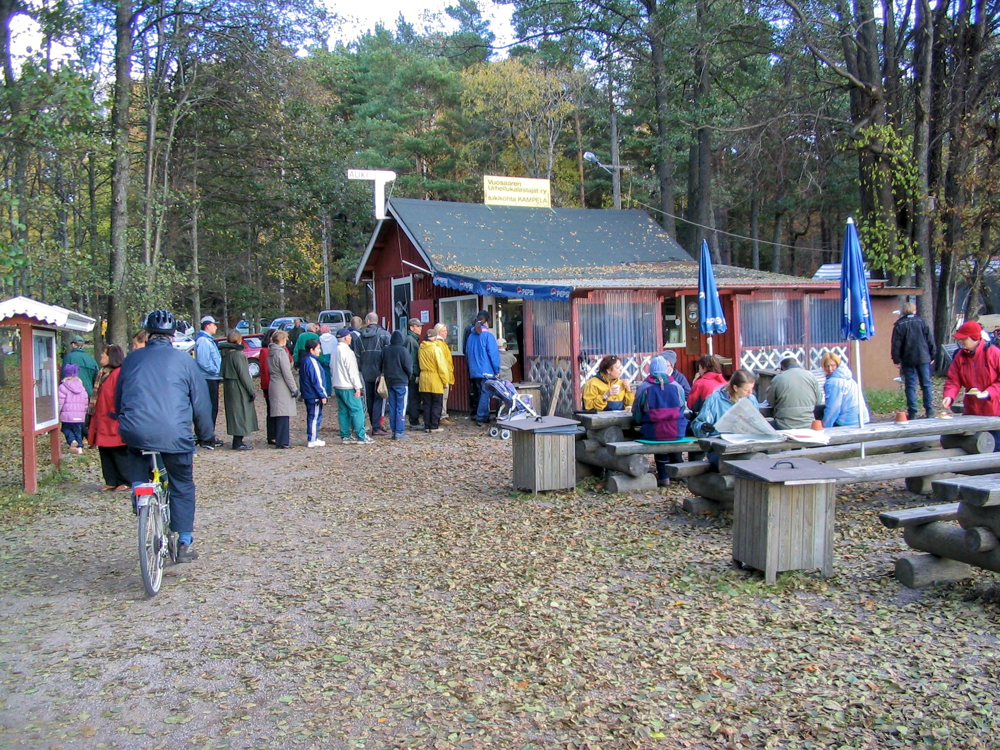 Kuvan keskellä pieni punainen kahvilamökki, jonne on ulkona kymmenen ihmisen jono. Kahvilassa on ulkokatos, jonka seinät on päällystetty läpinäkyvällä muovilla. Ulkona on hirsisiä penkkejä ja pöytiä, joiden ääressä ihmiset juovat kahvia, yksi lukee sanomalehteä. Hiekkakäytävä on pudonneiden lehtien peitossa. Vasemmassa reunassa joku lukee ilmoitustaulua. Katonharjalla on kyltti "Vuosaaren urheilukakalastajat ry tukikohta Kampela" ja katon päädyssä semaforimainen merkki "Auki". Taustalla mäntyjä ja syksyisiä tervaleppiä.
