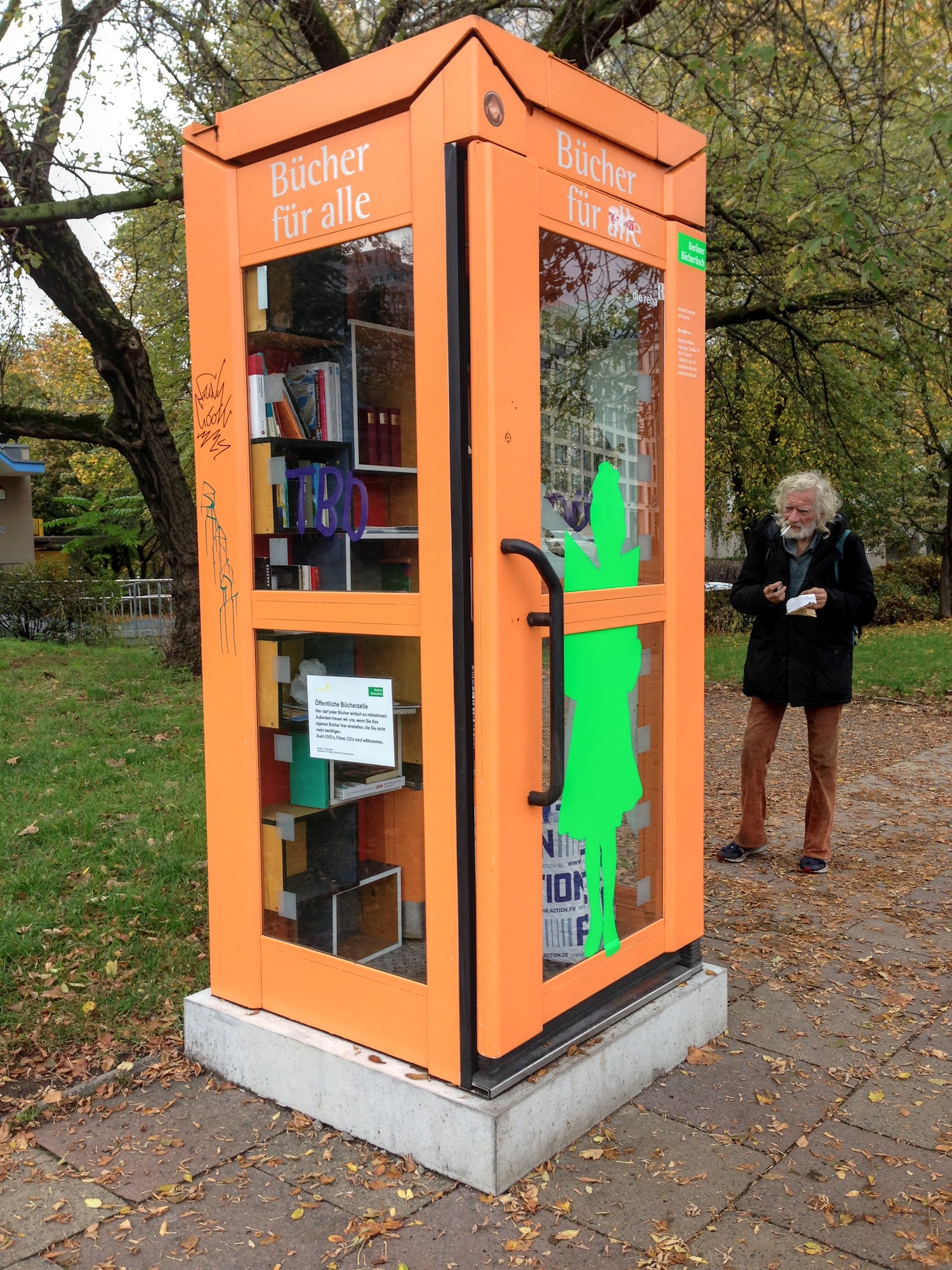 Jalkakäytävän reunassa puistikon laidassa on oranssi puhelinkoppi, jossa on sisällä kirjoja hyllyissä (ei puhelinta). Kopin yläreunassa on teksti "Bücher für alle". Oikealla kopista muutaman metrin kauempana on parrakas mies, joka pitää irtotupakkapussia kädessään ja on sytyttämässä savuketta.