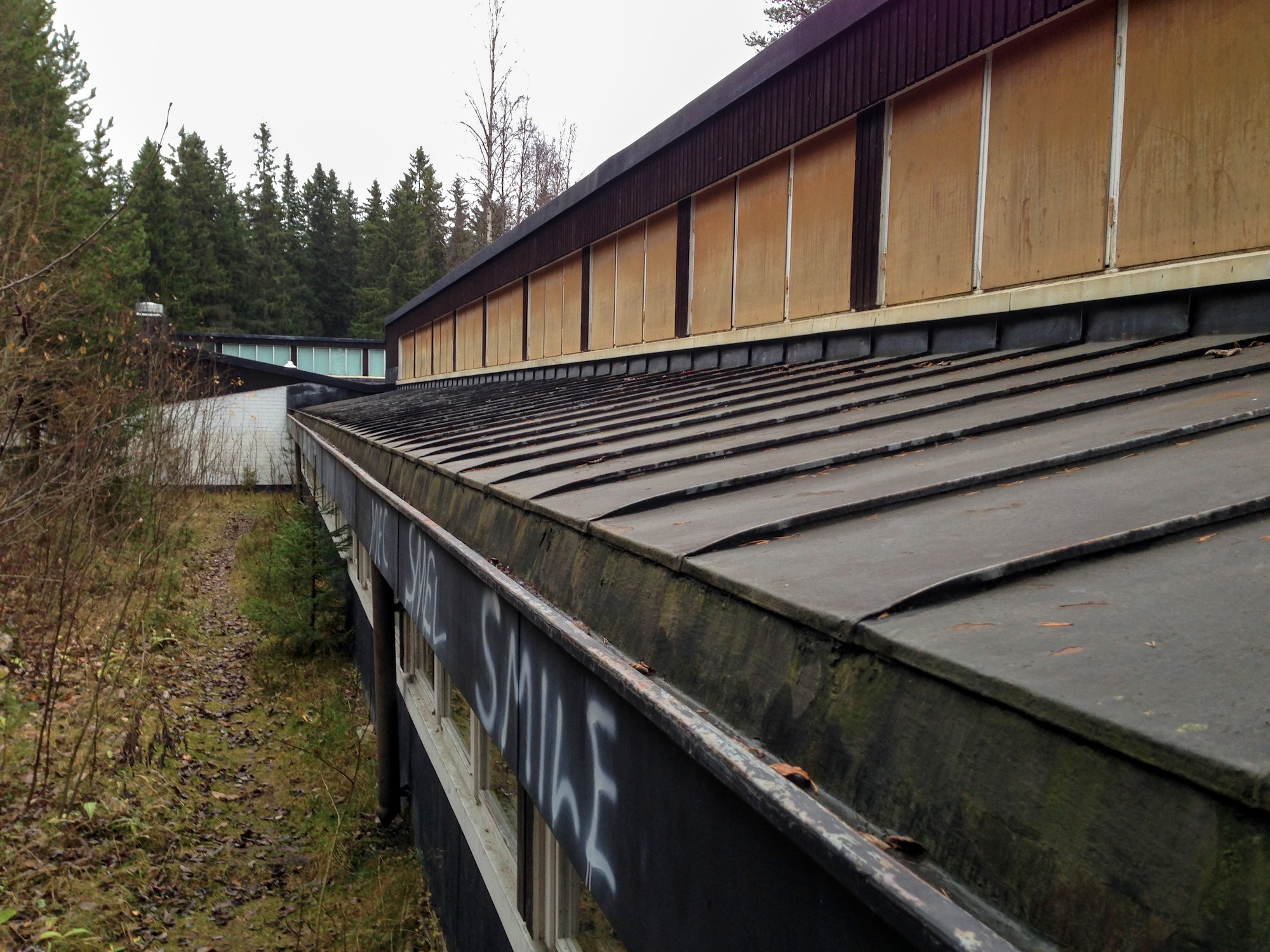 Syksyisessä metsänreunassa on matala koulurakennus. Kuva on otettu pitkittäin viiston peltikaton räystästä pitkin. Räystään alla käytävän ikkunarivi. Kapean katon toisella puolella olevan kohotetun osan ikkunat on peitetty vanerilla. Kauempana rakennus kääntyy suorakulmaisesti oikealle ja sen korkean osan ikkunat näkyvät kuvan ottajan suuntaan. Räystääseen on suihkemaalilla kirjoitettu "Smile".