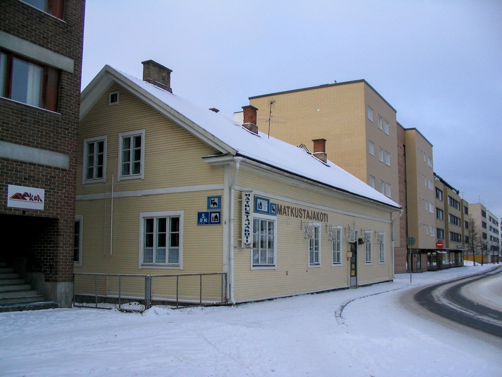 Talvisen kadun varrella uudehkojen nelikerroksisten talojen välissä on kaksikerroksinen puutalo, matkustajakoti. Talon reuna on useita metrejä kerrostalojen seinälinjaa ulompana.