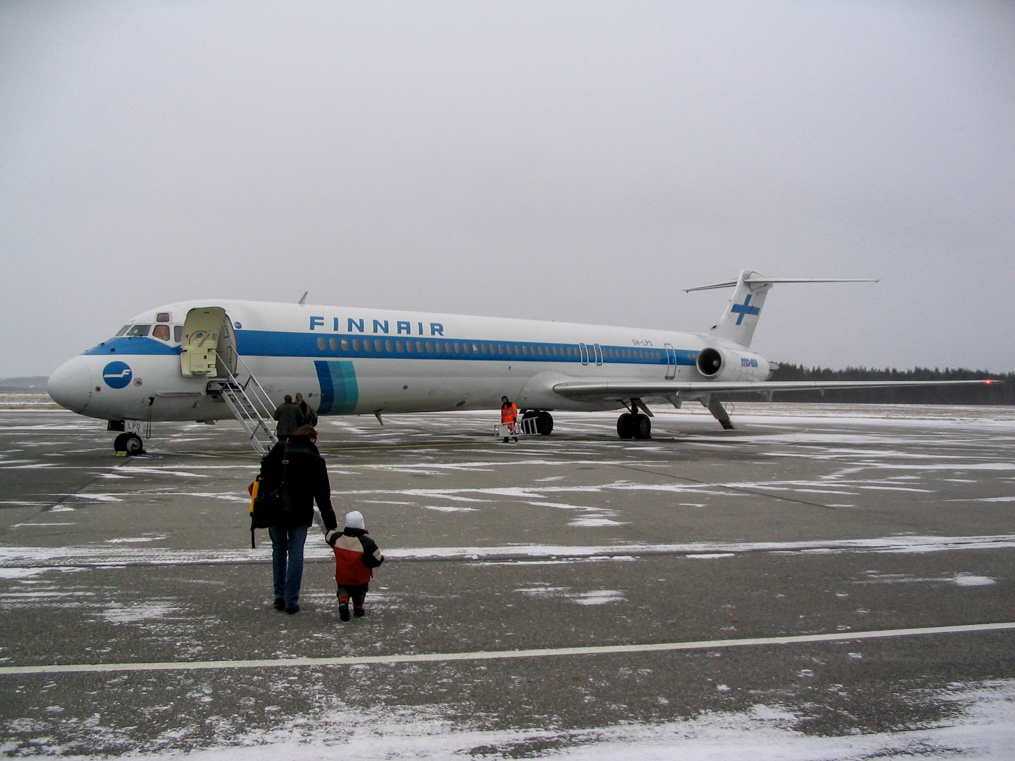 Matkustajakone (Finnairin MD-83) lentoaseman asfaltilla. Matkustajia kävelee kohti koneesta laskeutuvia portaita, etualalla nainen ja pieni lapsi käsi kädessä. Työntekijä kantaa tikkaita koneen luona siiven kohdalla. Maassa on lunta, asfalttitasollakin laikuittain.