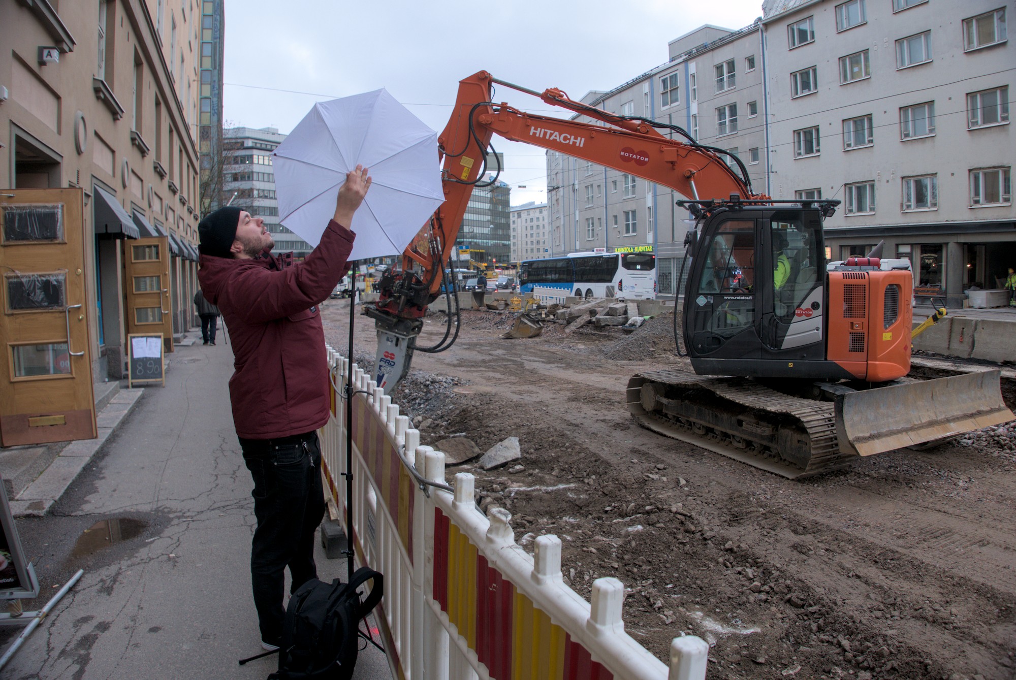 Valokuvaaja pystyttää valaisimen sateenvarjomaista varjostinta jalkakäytävän reunassa katutyömaan vieressä. Työmaa-aidan oikealla puolella remontissa olevalla ajorata-alueella kone muokkaa maata.